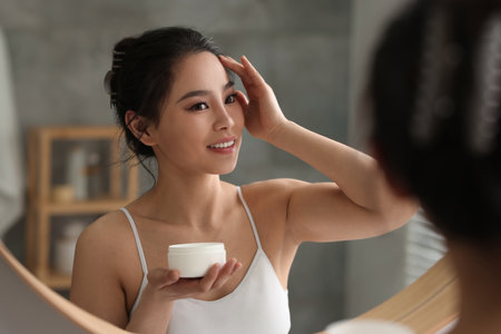 Happy woman applying face cream near mirror at homeの写真素材