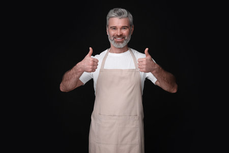 Happy man in kitchen apron showing thumbs up on black background. Mockup for designの写真素材