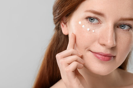 Beautiful woman with freckles and cream on her face against grey background, closeup. Space for textの写真素材