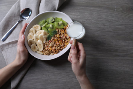 Woman pouring milk into bowl of tasty granola with banana and kiwi at gray wooden table, top viewの写真素材