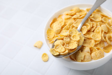 Breakfast cereal. Tasty corn flakes with milk in bowl and spoon on white tiled table, closeup. Space...
