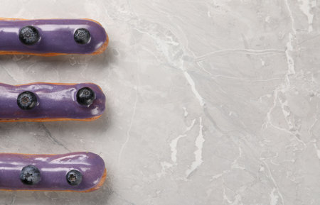 Tasty glazed eclairs with blueberries on gray marble table, flat lay. Space for text