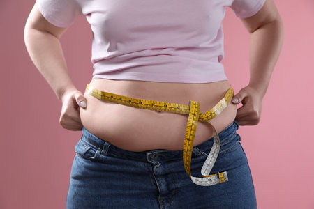 Woman with measuring tape touching belly fat on pink background, closeup. Overweight problemの写真素材