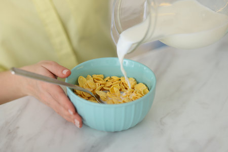Making breakfast. Woman pouring milk from jug into bowl with cornflakes at white marble table,...