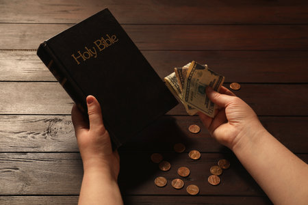 Donate and give concept. Woman with dollar banknotes, coins and Bible at wooden table, closeupの写真素材