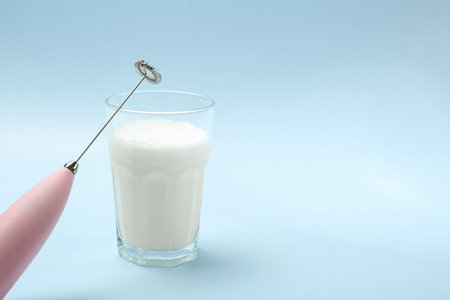 Mini mixer (milk frother) and whipped milk in glass on light blue background. Space for textの写真素材