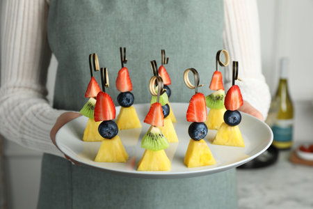 Woman holding plate of tasty canapes with pineapple, kiwi and berries indoors, closeupの写真素材