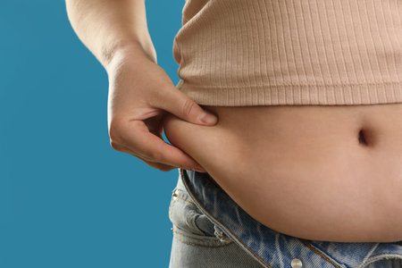 Woman touching belly fat on light blue background, closeup. Overweight problem