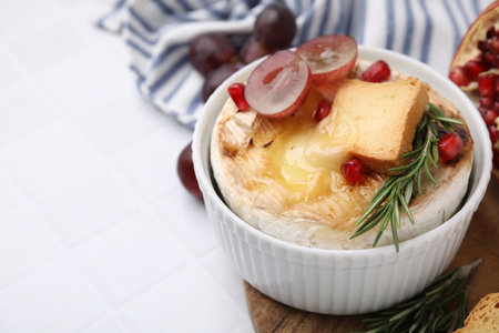 Tasty baked camembert with crouton, grape and rosemary on white tiled table, closeup. Space for textの写真素材