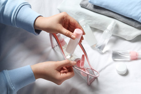 Cosmetic travel kit. Woman putting small bottle with personal care product into plastic bag on bed,...