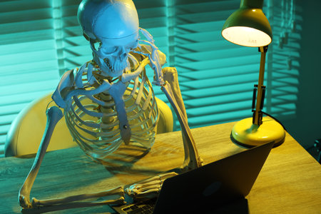 Waiting concept. Human skeleton sitting at wooden table with laptop indoors