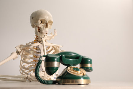Waiting concept. Human skeleton at table with corded telephone against light gray background, space...