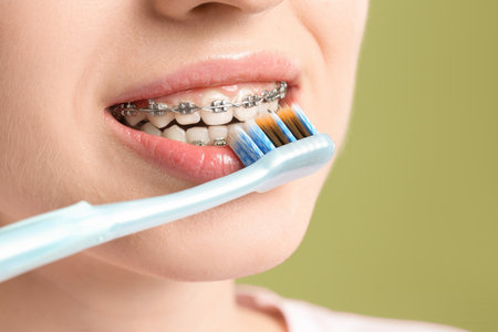 Smiling woman with dental braces cleaning teeth on light green background, closeup