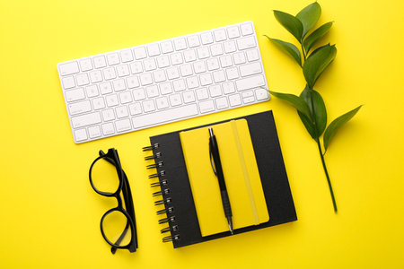 Office stationery, glasses, computer keyboard and green leaves on yellow background, flat layの写真素材