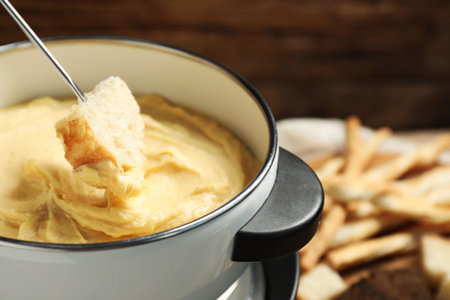 Dipping piece of bread into fondue pot with melted cheese on blurred background, closeupの写真素材