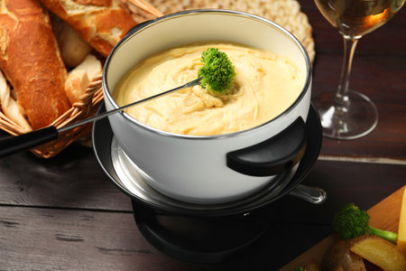 Fork with piece of broccoli, melted cheese in fondue pot and snacks on wooden table, closeupの写真素材