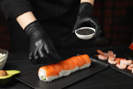 Chef in gloves adding sesame seeds onto tasty sushi rolls at black wooden table, closeupの写真素材