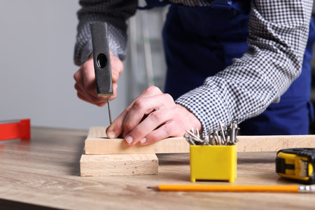 Professional repairman hammering nail into board at wooden table indoors, closeupの写真素材