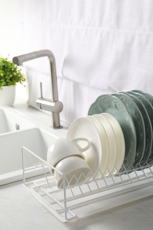 Drainer with different clean dishware and cups on white table in kitchenの写真素材