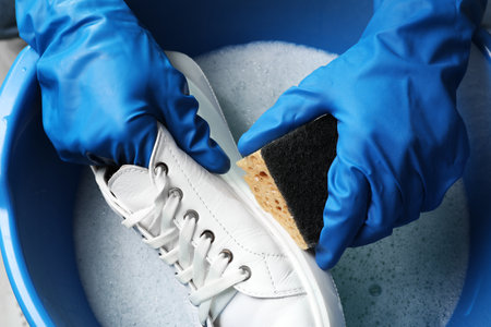 Woman with gloves and sponge cleaning stylish sneakers in wash basin, top view