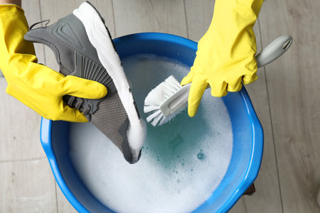 Woman with gloves and cleaning brush stylish sneakers in wash basin, top view