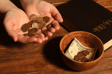 Donate and give concept. Woman holding coins, closeup. Bible and bowl of money on wooden table
