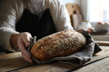Man holding loaf of fresh bread at wooden table indoors, closeup