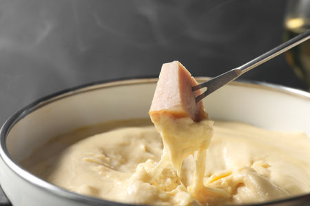 Dipping piece of ham into fondue pot with melted cheese on gray background, closeupの写真素材