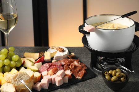 Fork with piece of ham, fondue pot with melted cheese, wine and snacks on gray tableの写真素材