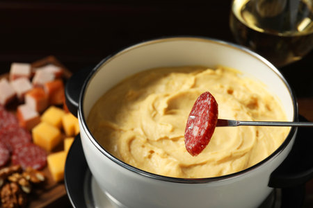 Dipping piece of sausage into fondue pot with melted cheese at table, closeupの写真素材