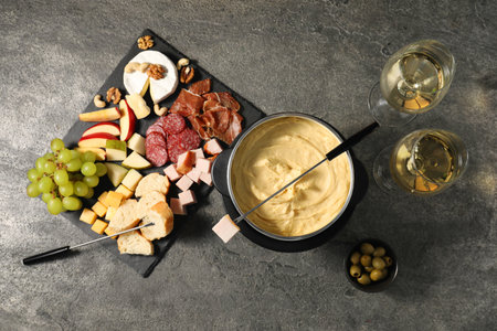 Fork with piece of ham, fondue pot with melted cheese, wine and snacks on gray textured table, flat layの写真素材