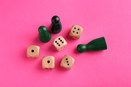 Wooden dices and color game pieces on pink background, flat lay