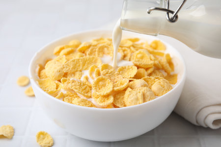 Breakfast cereal. Pouring milk into bowl with tasty corn flakes at white table, closeup