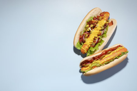 Delicious hot dogs with bacon, lettuce, onion and pickle on light blue background, flat lay. Space...
