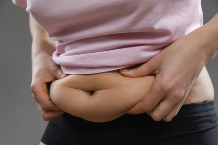 Woman touching belly fat on gray background, closeup. Overweight problem