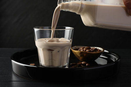 Pouring coffee cream liqueur into glass at black wooden table, closeup