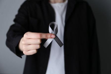 Woman holding gray awareness ribbon on color background, closeup