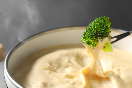 Dipping piece of broccoli into fondue pot with melted cheese on gray background, closeupの写真素材