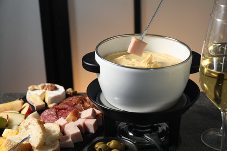 Dipping piece of ham into fondue pot with melted cheese at gray table with wine and snacks, closeupの写真素材