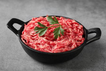 Raw ground meat and parsley in bowl on gray table, closeup