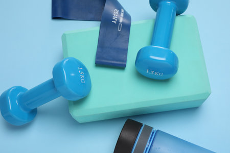 Two dumbbells, yoga block, fitness elastic band and bottle on light blue background, above view