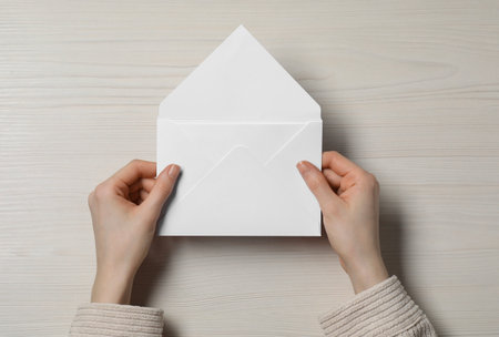 Woman holding letter envelope with card at light wooden table, top view