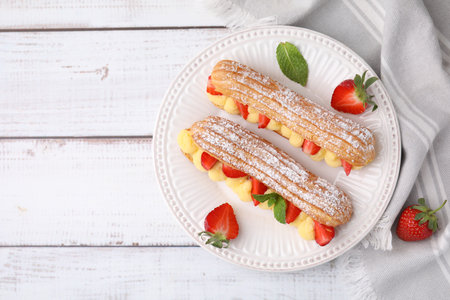 Delicious eclairs filled with cream, strawberries and mint on white wooden table, top view. Space...
