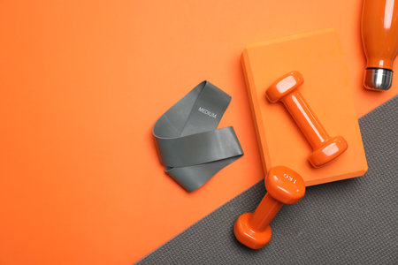 Dumbbells, yoga block, mat, thermo bottle and fitness elastic band on orange background, flat lay....