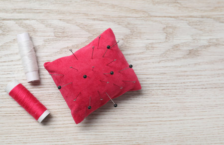 Red pincushion with sewing pins and spools of threads on light wooden table, flat lay. Space for...