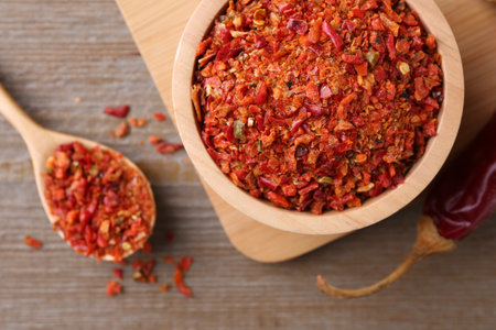 Aromatic spice. Red chili pepper flakes in bowl, spoon and pod on wooden table, flat lay