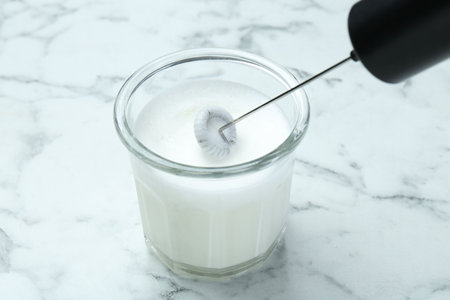 Whisking milk in glass with mini mixer (milk frother) at white marble table, closeupの写真素材