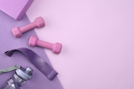 Two dumbbells, yoga block, mat, fitness elastic band and bottle on violet background, flat lay. Space for textの写真素材