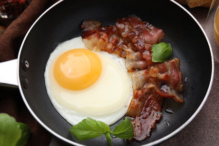 Fried egg, bacon and basil in frying pan on brown tableの写真素材