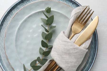 Stylish setting with cutlery, napkin, eucalyptus branch and plates on white table, top view